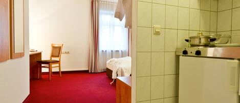 Habitación de hotel con alfombra roja, una silla de madera, una cama, un escritorio pequeño y una kitchenette con estufa y refrigerador.