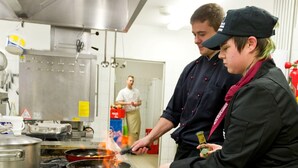 Una cocina con electrodomésticos de acero inoxidable, una persona cocinando en la estufa y otra sosteniendo una botella.