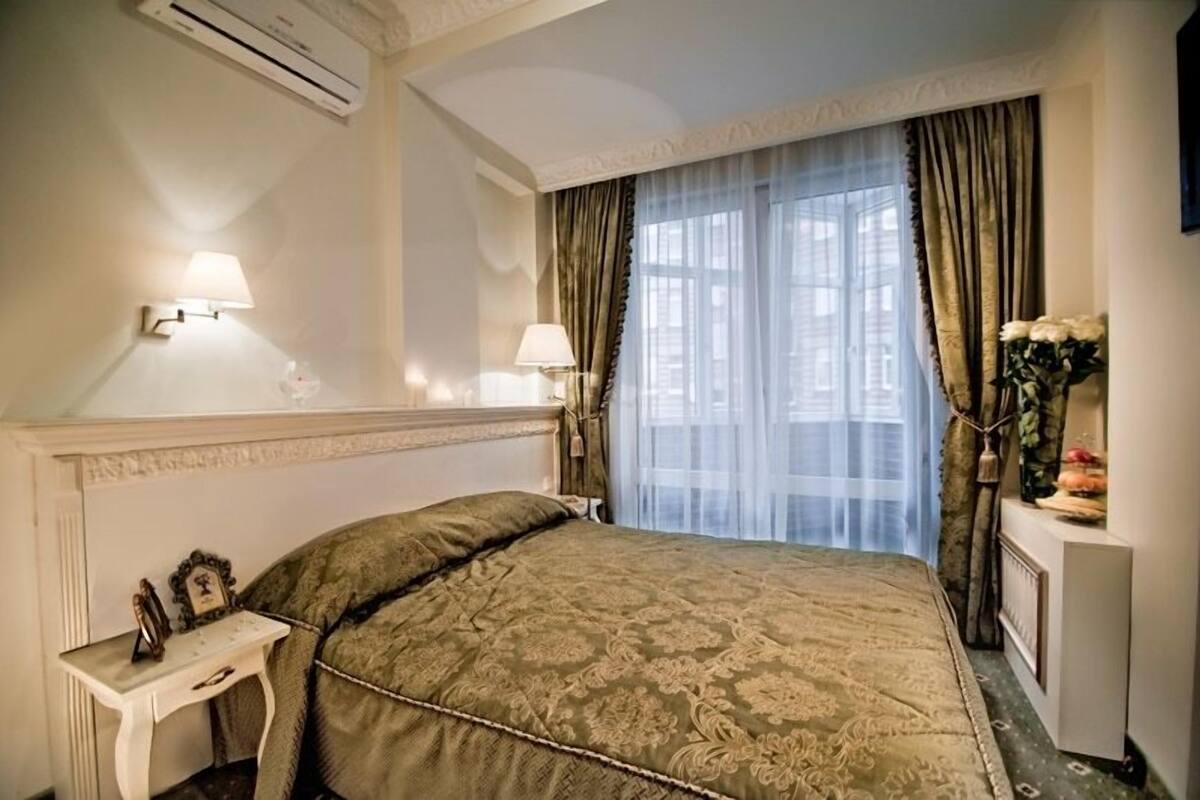 A hotel room with a large bed, two bedside lamps, a nightstand with a lamp, a dresser with a vase of flowers, and a window with sheer curtains.