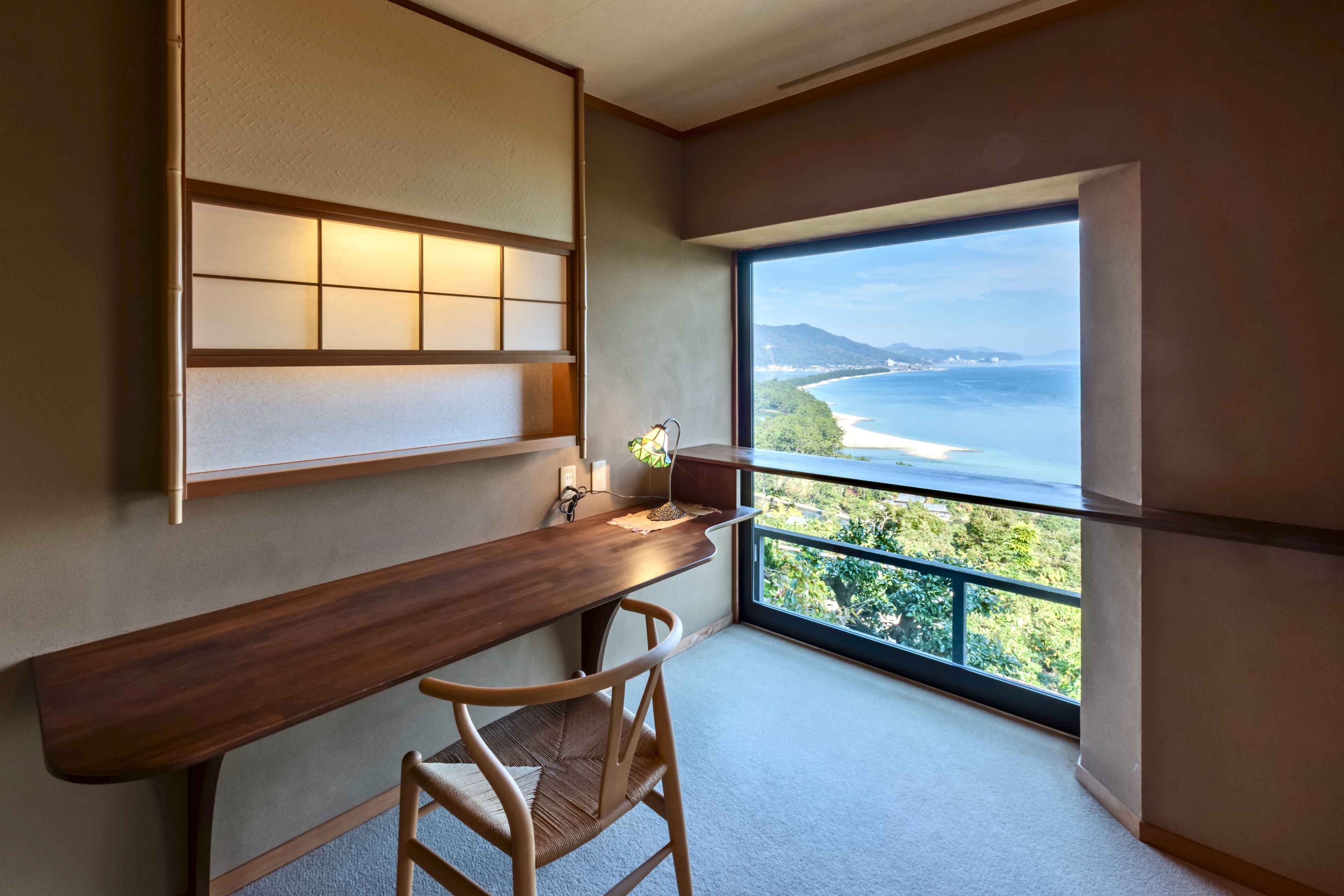 A room with a wooden desk, a chair, a large window with a view of the sea and greenery, and a wall-mounted shelf with lights.