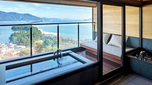 A modern bathroom with a large window, a built-in sink, and a seating area with a view of a coastal landscape. - Genmyoan (Miyazu)