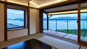 A room with tatami flooring, a sliding glass door to a balcony with a view of a body of water and mountains. - Genmyoan (Miyazu)