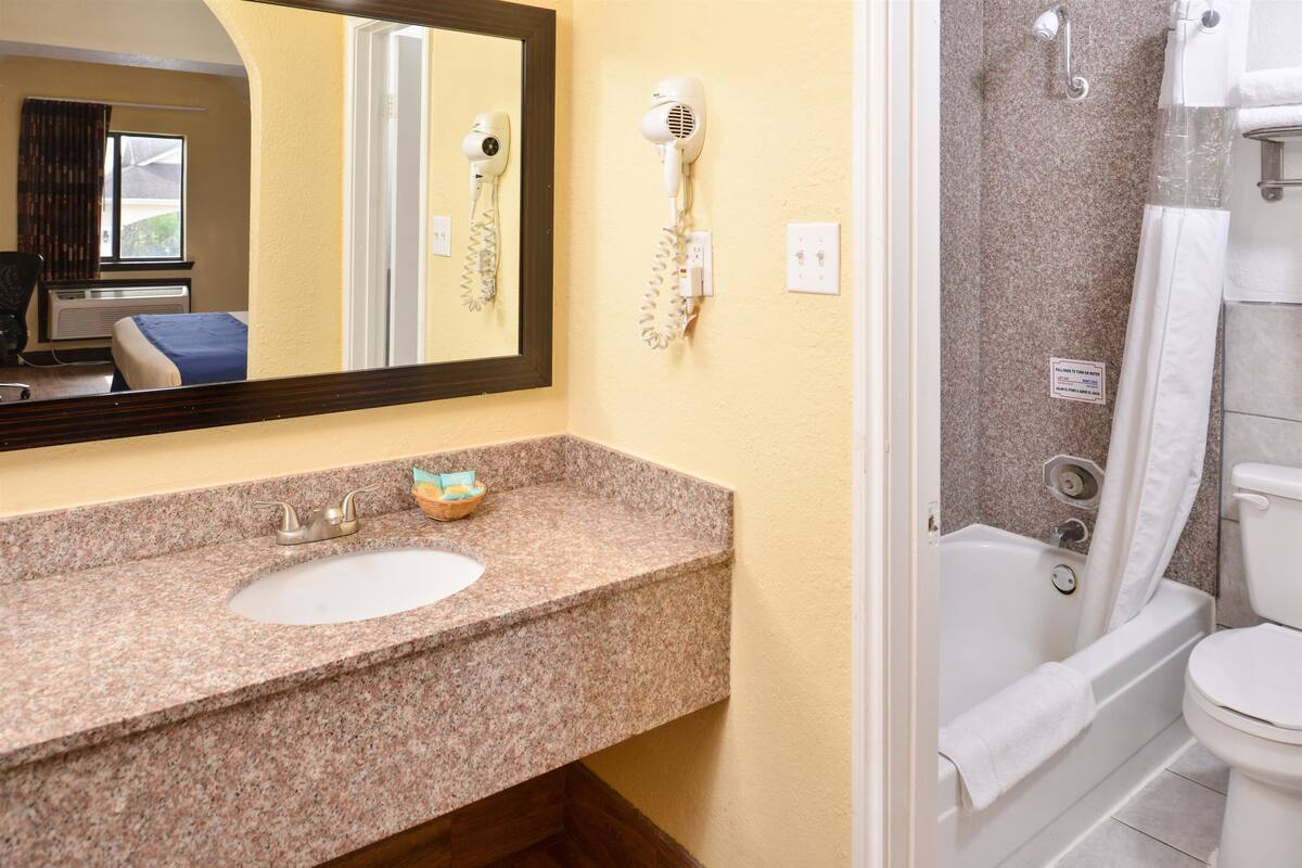 A bathroom with a granite countertop sink, a hairdryer mounted on the wall, a bathtub, and a toilet.