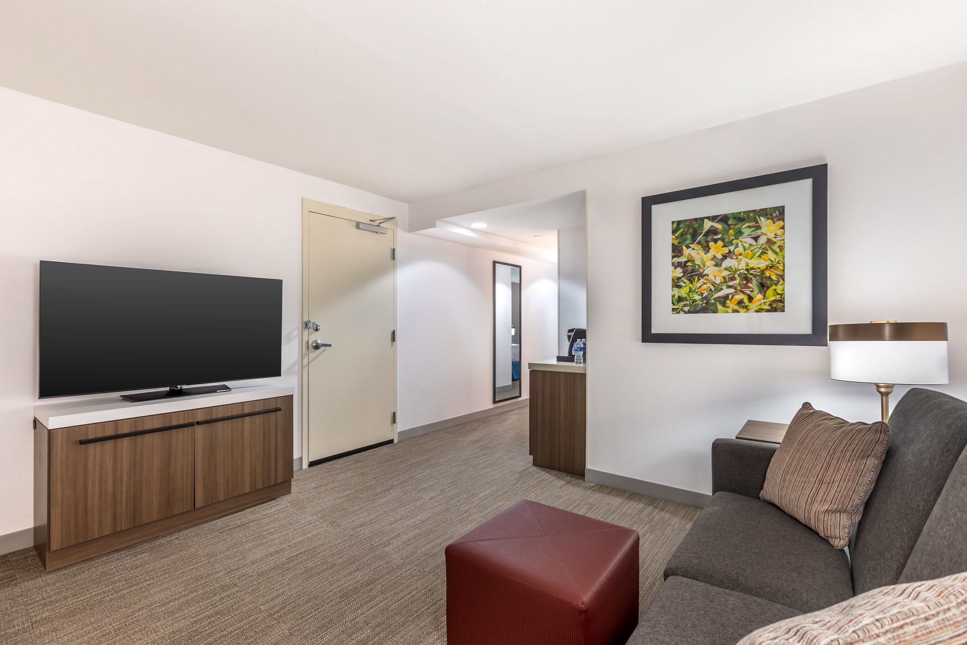 A hotel room with a flat-screen TV, a sofa, a red ottoman, a framed picture, and a lamp.