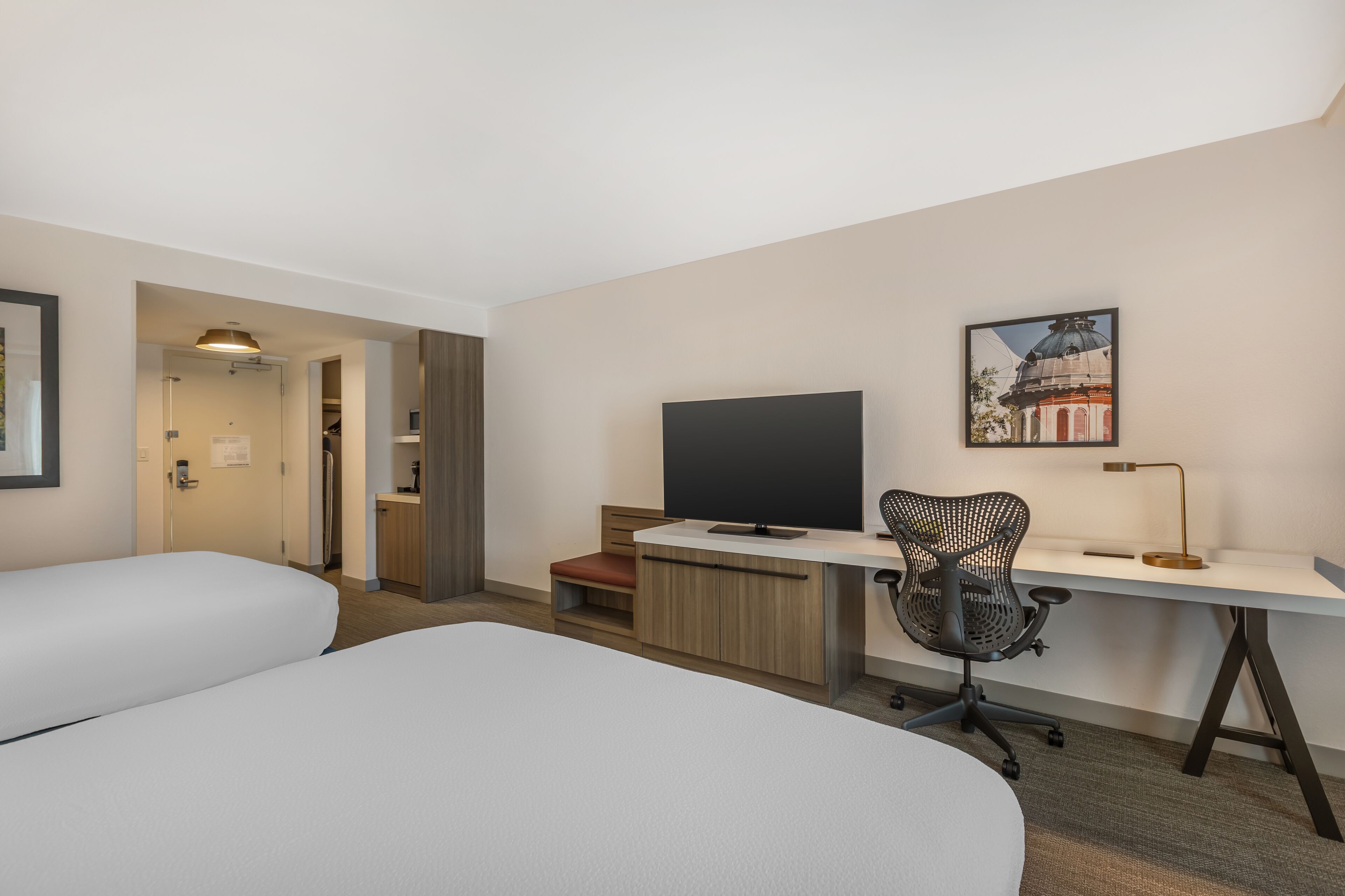 A hotel room with two beds, a desk with a computer, a chair, a desk lamp, and a framed picture on the wall.