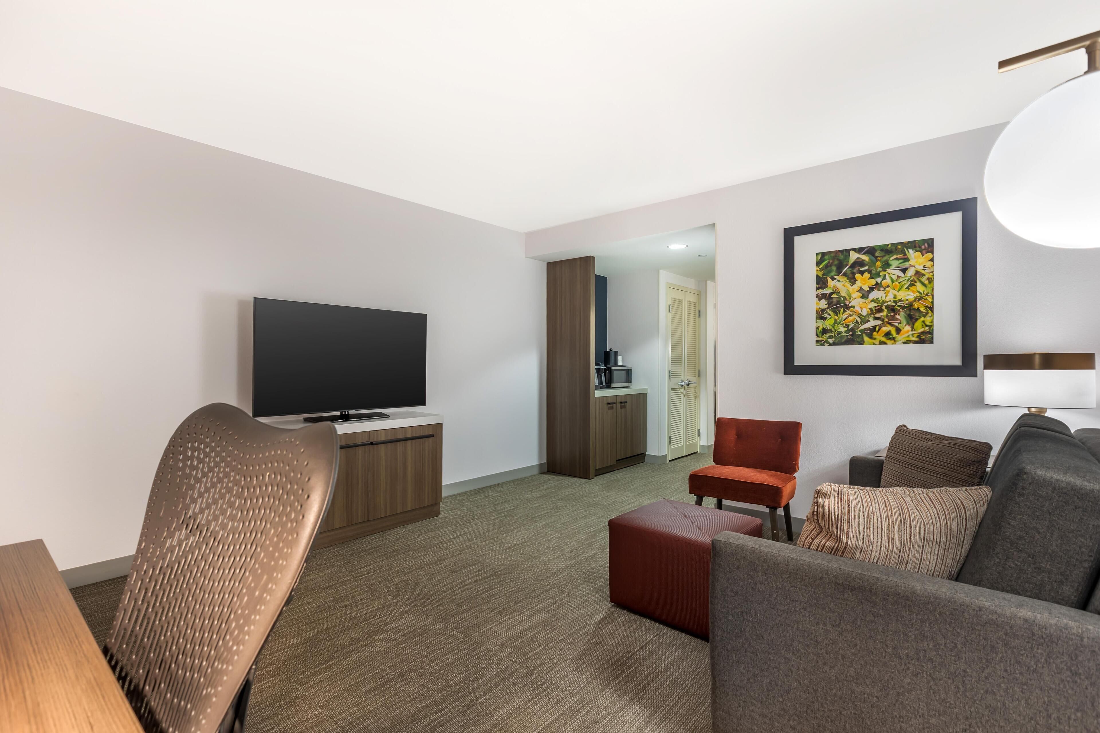 Ett modernt hotellrum med en platt-TV, en soffa, ett matbord och en röd fåtölj.