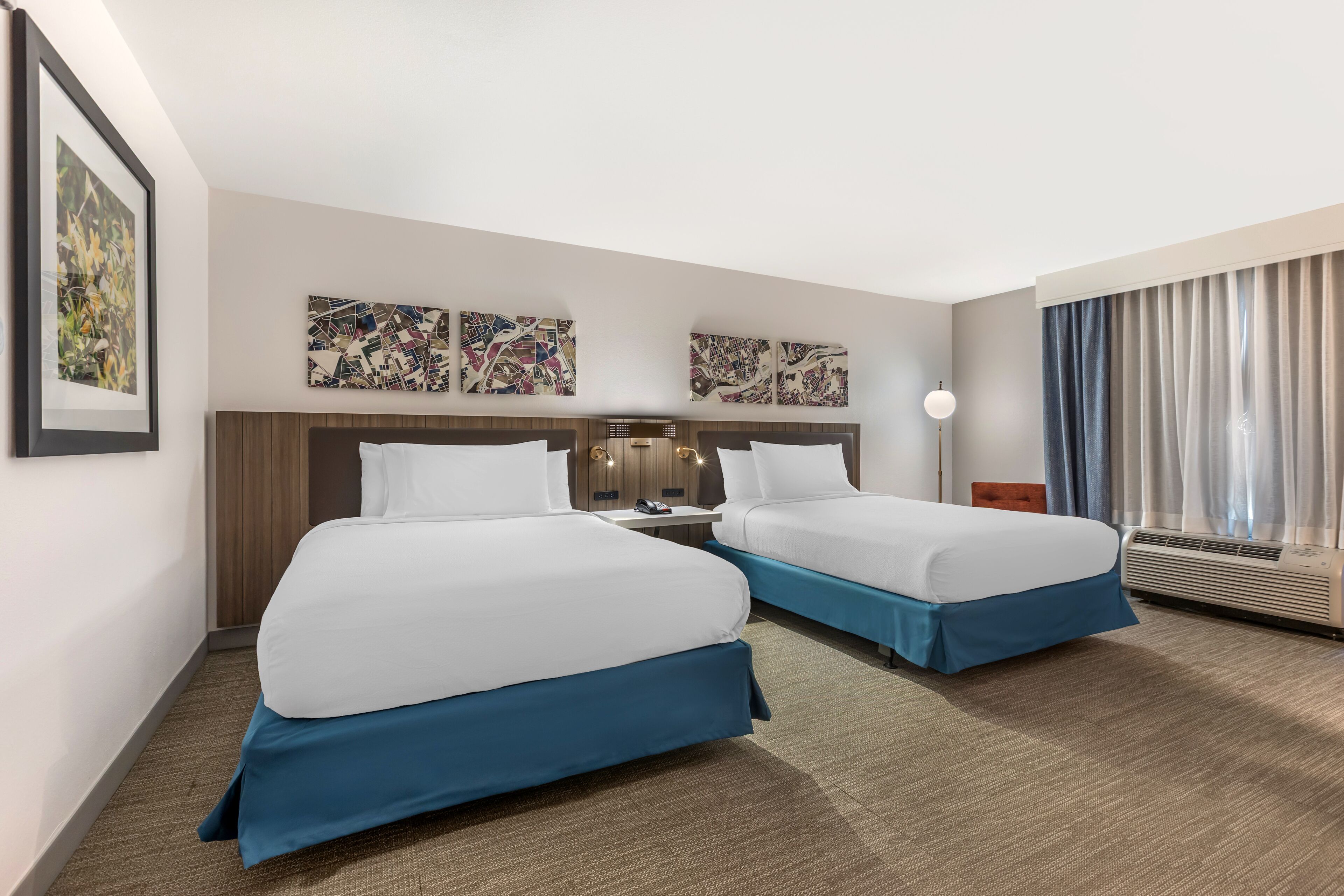 A hotel room with two beds, a large window with curtains, and artwork on the wall.