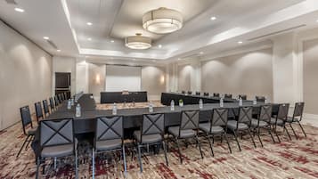 Un salón de conferencias con una larga mesa, sillas, una pantalla de proyector y una computadora portátil.