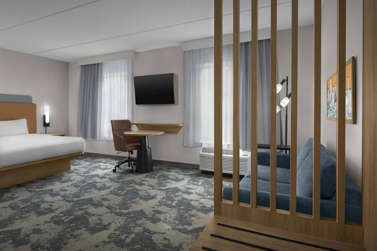 A hotel room with a bed, a desk, a chair, a TV, a sofa, and a large window with curtains.