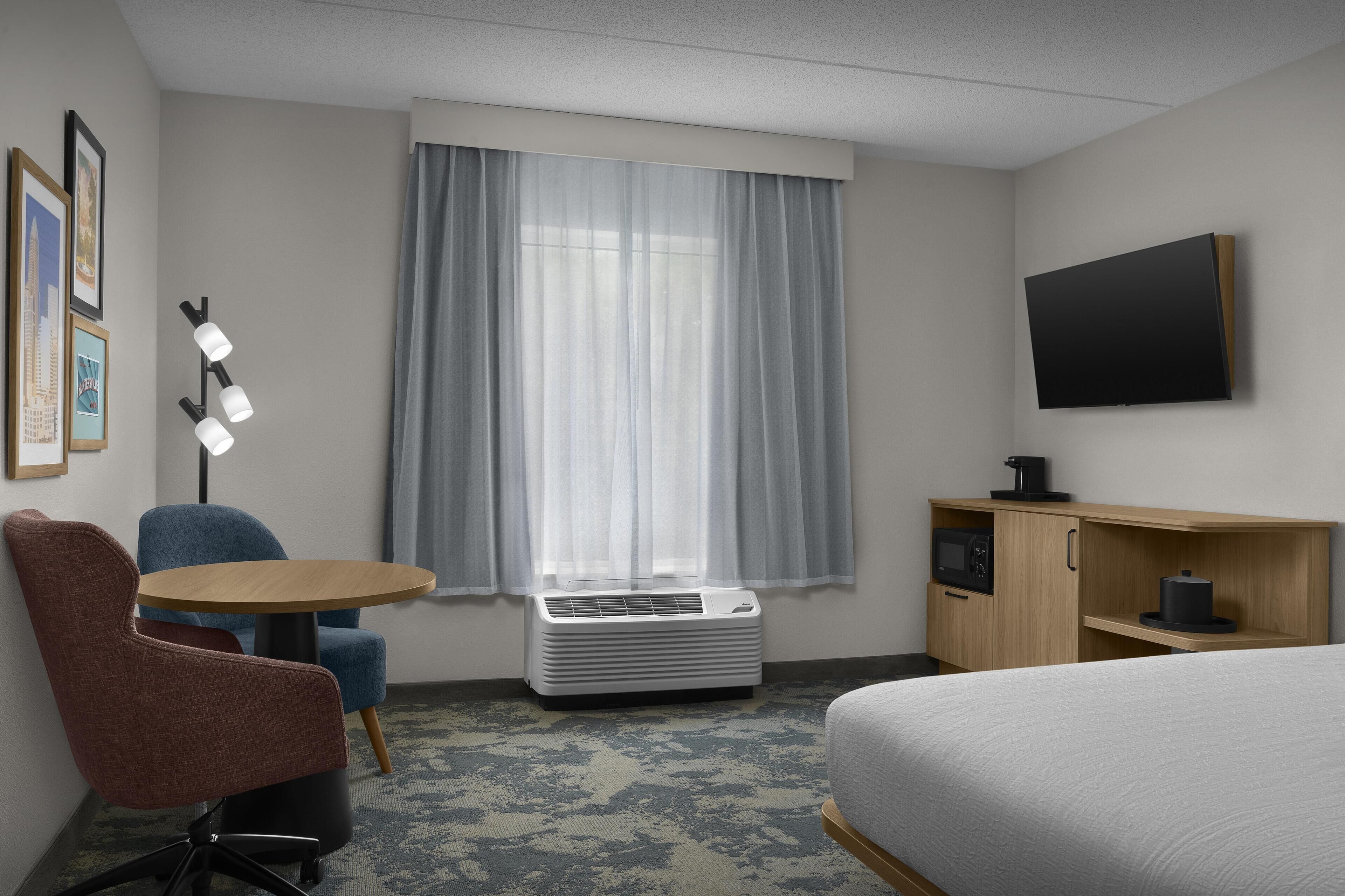 A hotel room with a bed, a desk with a chair, a flat-screen TV, an air conditioner, and a window with curtains.