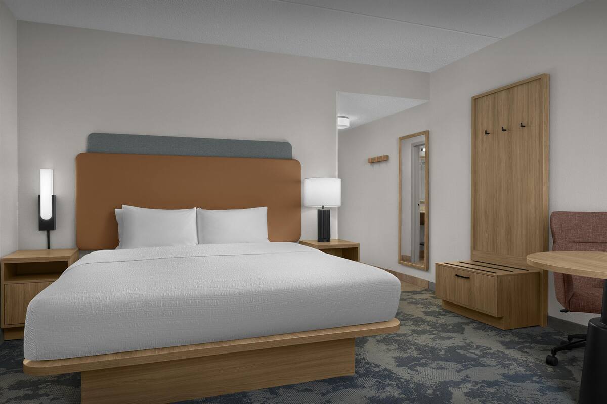 A hotel room with a large bed, bedside tables, a desk, a chair, a wardrobe, and a mirror.