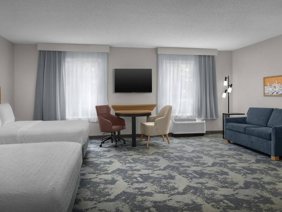 A hotel room with two beds, a desk, two chairs, a sofa, a flat-screen TV, and a painting on the wall.