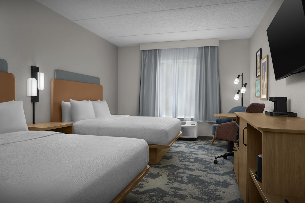 A hotel room with two beds, a desk, a chair, a TV, and a window with curtains.