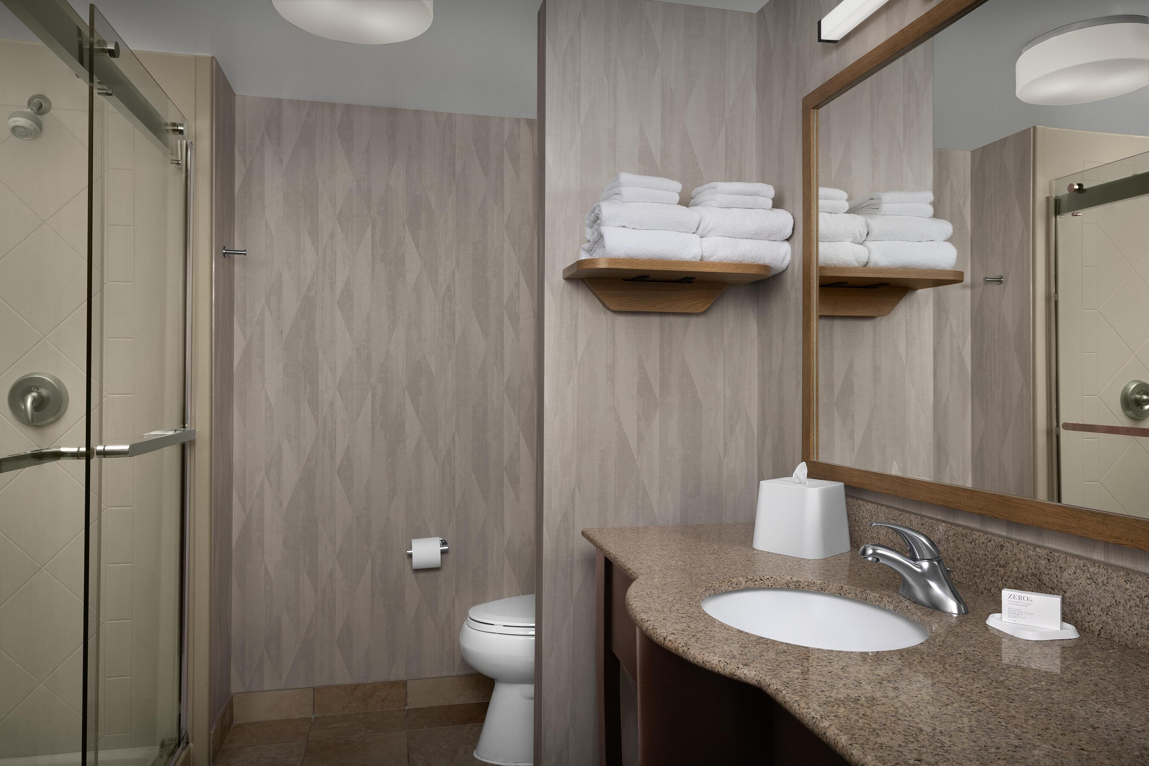 Une salle de bain dotée d’une douche à parois de verre, de toilettes, d’un lavabo avec un plan de travail en granit et d’un miroir au-dessus duquel se trouve un porte-serviettes.