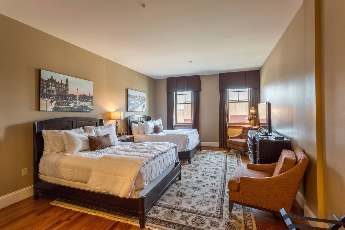 A hotel room with two beds, a desk, a chair, and a large window.