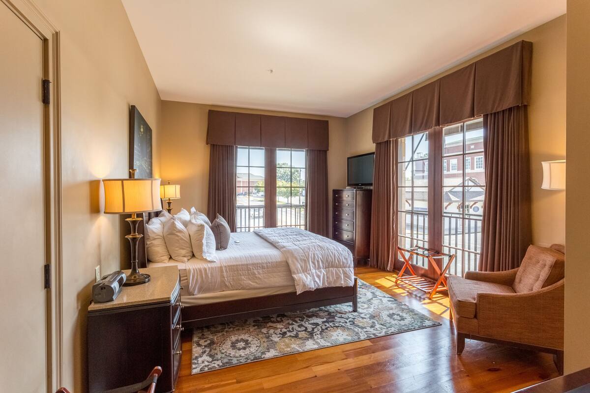 A hotel room with a large bed, a desk with a lamp, a chair, and a view of the outside through large windows.