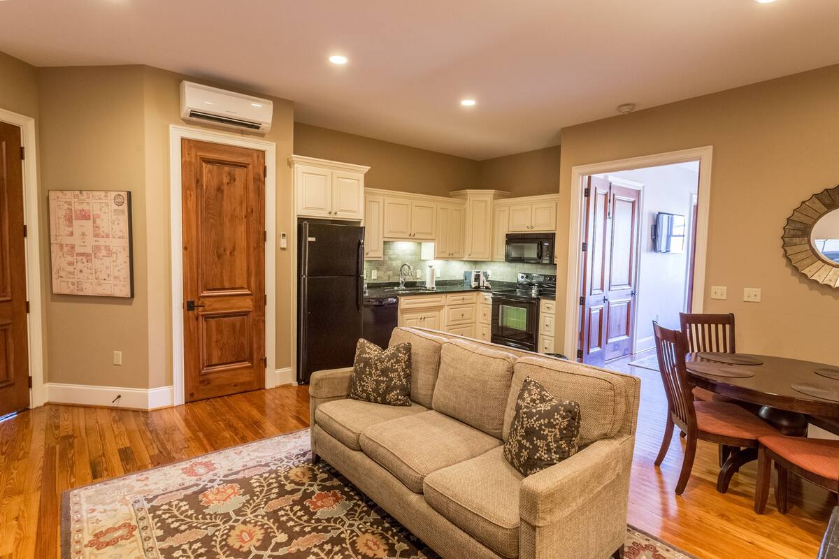 A living room with a beige sofa, a patterned rug, wooden flooring, and a kitchen in the background.
