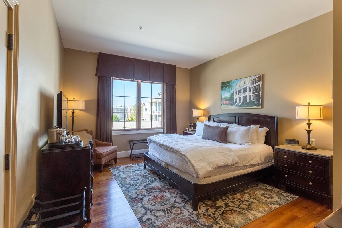 A bedroom with a large bed, two bedside tables with lamps, a dresser, a chair, and a window with curtains.