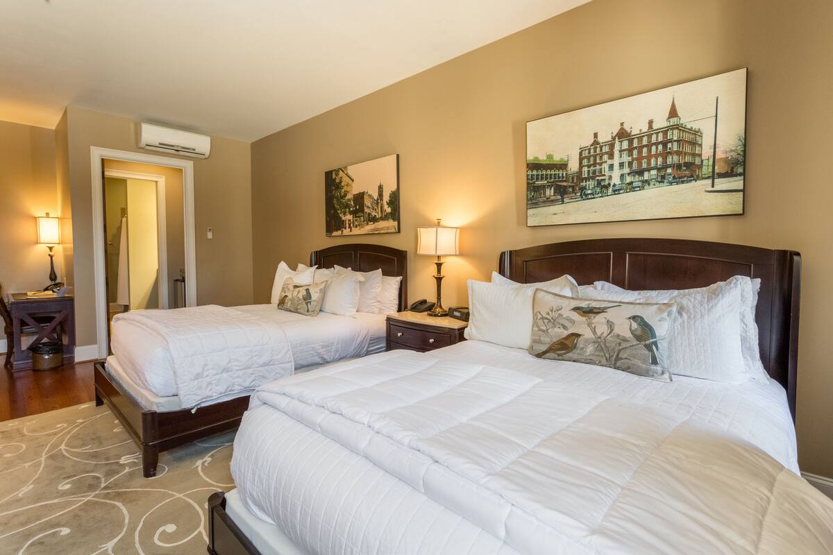 A hotel room with two beds, a wooden headboard, a beige wall, and a framed cityscape picture.