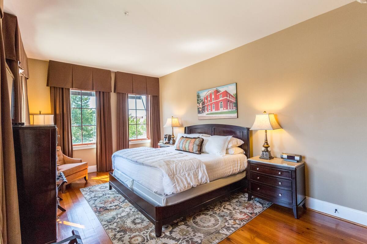 A bedroom with a large bed, a nightstand, a dresser, a chair, and a window with curtains.