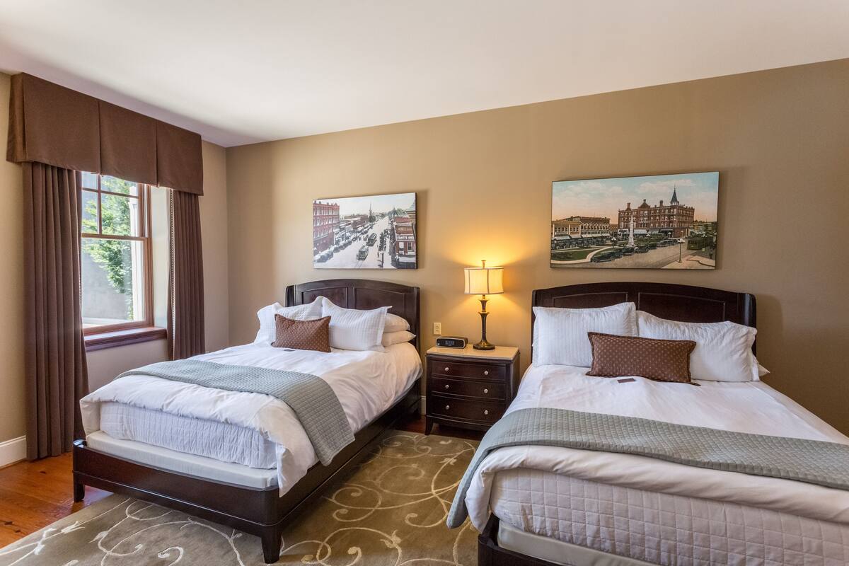 A hotel room with two beds, a window with curtains, a nightstand with a lamp, and two wall-mounted paintings.