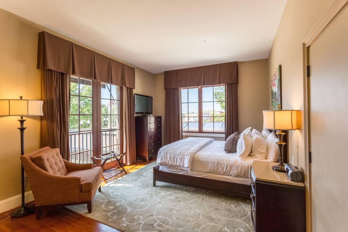 A hotel room with a bed, a chair, a desk, a television, and large windows.