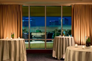 Una habitación con dos mesas redondas, cada una con una vela y un recipiente de vidrio. Se ven ventanas amplias con cortinas, que ofrecen vistas de edificios y del paisaje urbano de noche.