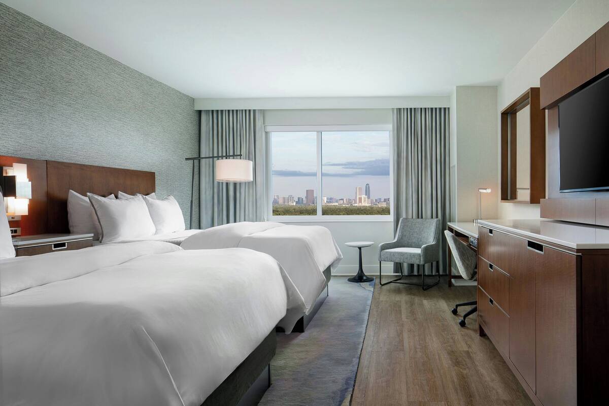A hotel room with two beds, a desk, a chair, a TV, and a large window with a city view.