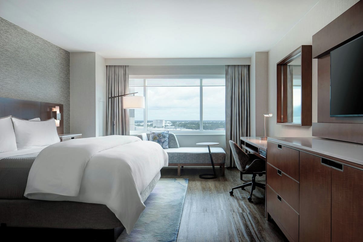 A hotel room with a large bed, a desk, a chair, a television, and a view of the city.