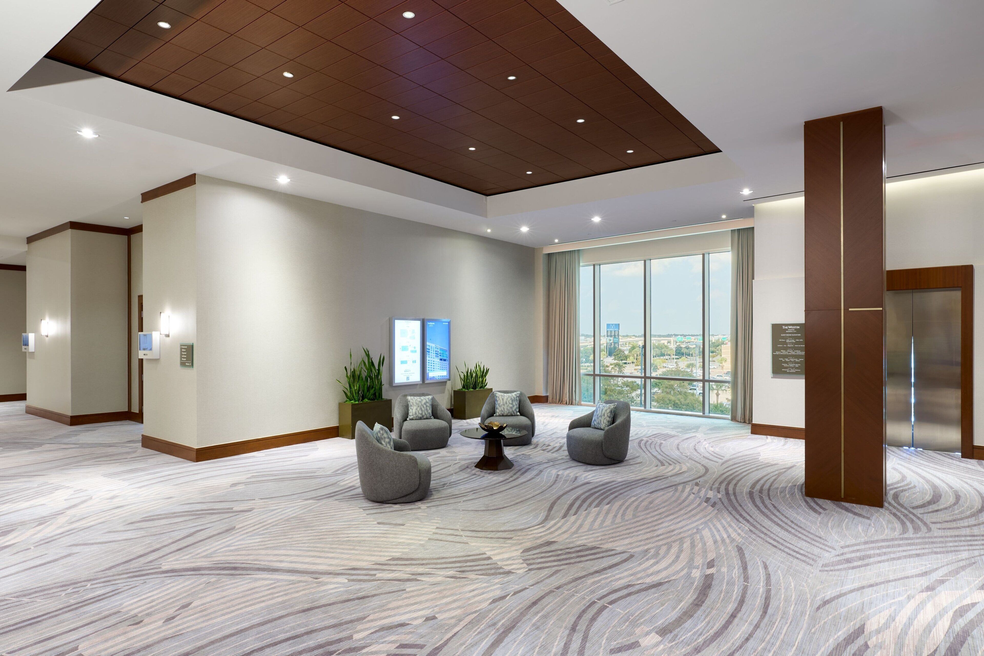 A modern lobby with a marble floor, grey armchairs, a central coffee table, and a large window offering a city view.