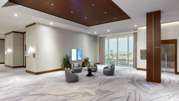 A modern lobby with a marble floor, grey armchairs, a central coffee table, and a large window offering a city view.