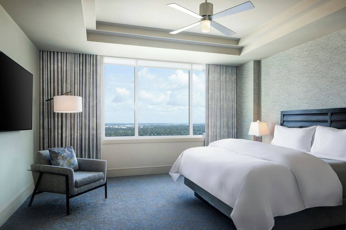 A modern hotel room with a large bed, a chair, a ceiling fan, a window with a view, and a flat-screen TV.