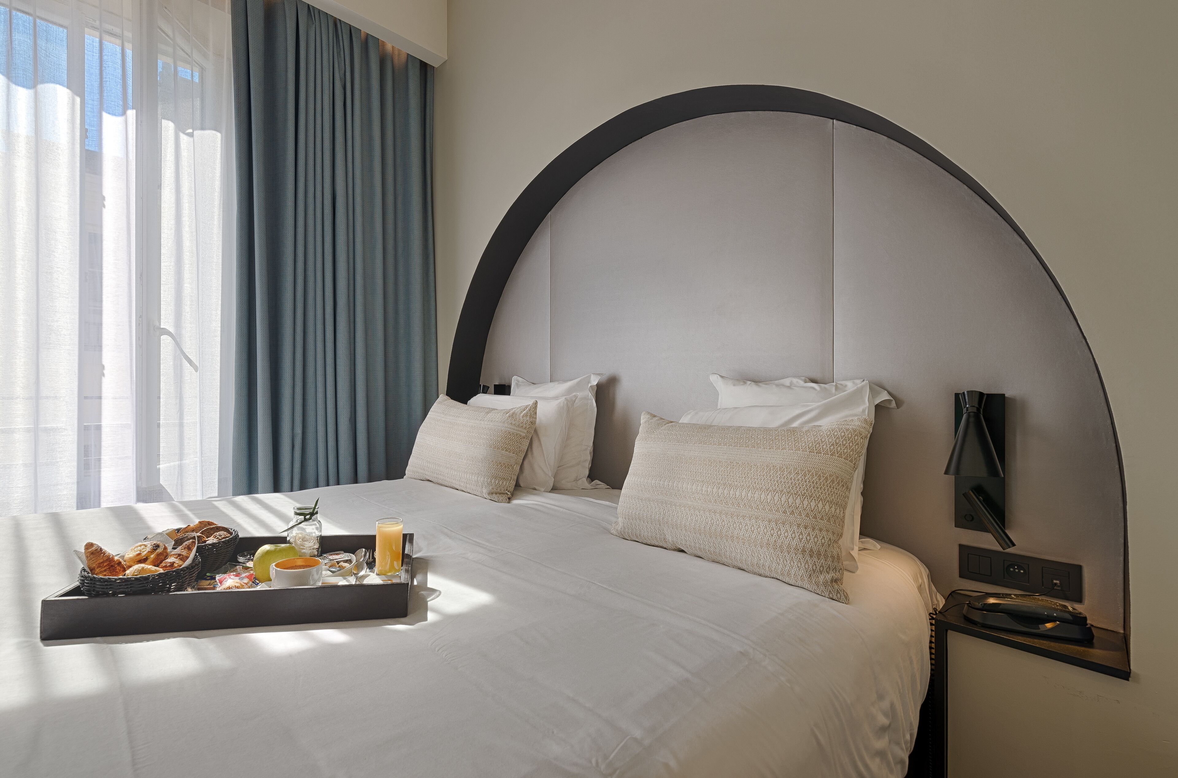Habitación de hotel con una cama grande, una bandeja con alimentos para el desayuno, una mesita de noche con lámpara y vistas al exterior.
