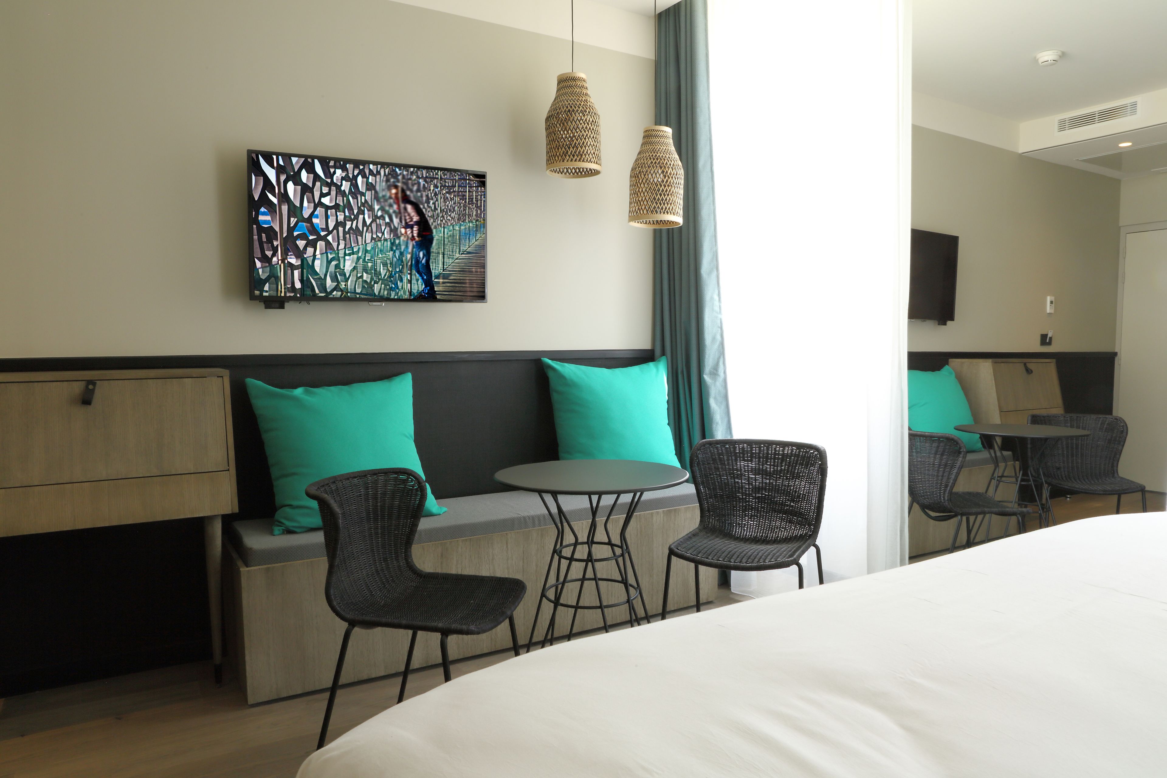 Een hotelkamer met een bed, twee stoelen, een klein tafeltje, een bank met turquoise kussens, een aan de muur bevestigde televisie en twee hangende rieten lampen.