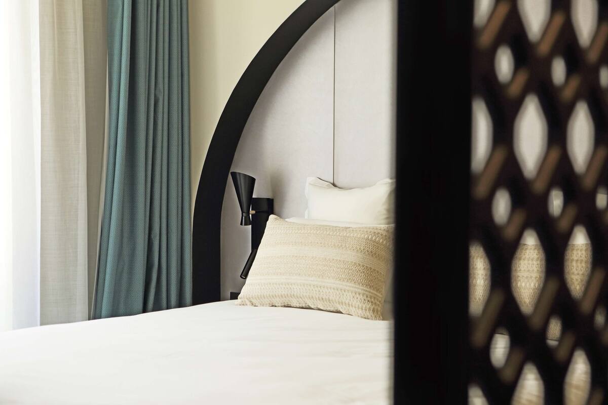 Een netjes opgemaakt bed met een witte deken en een crèmekleurig kussen, een zwarte lamp op een nachtkastje, en een blauwgroen gordijn dat deels opzij getrokken is.