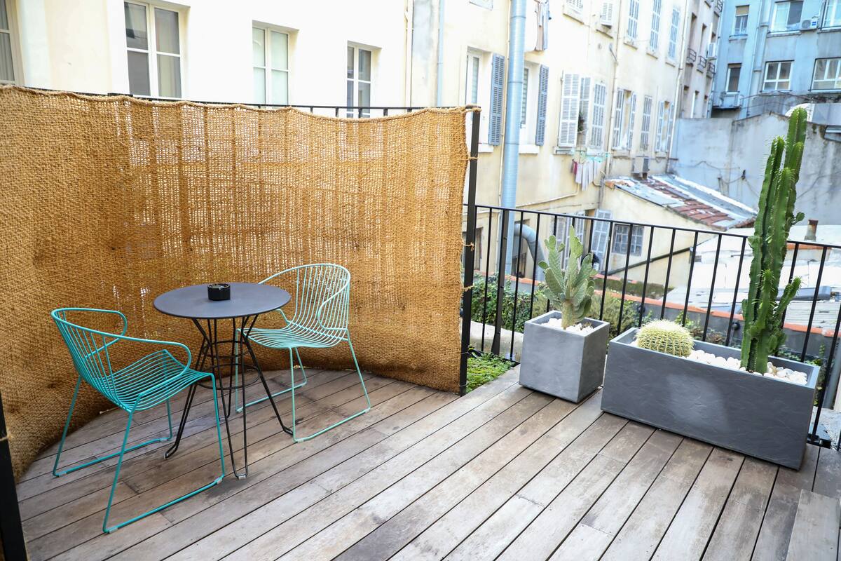 Een balkon met een geweven scherm, twee metalen stoelen, een kleine tafel en potten met cactussen.