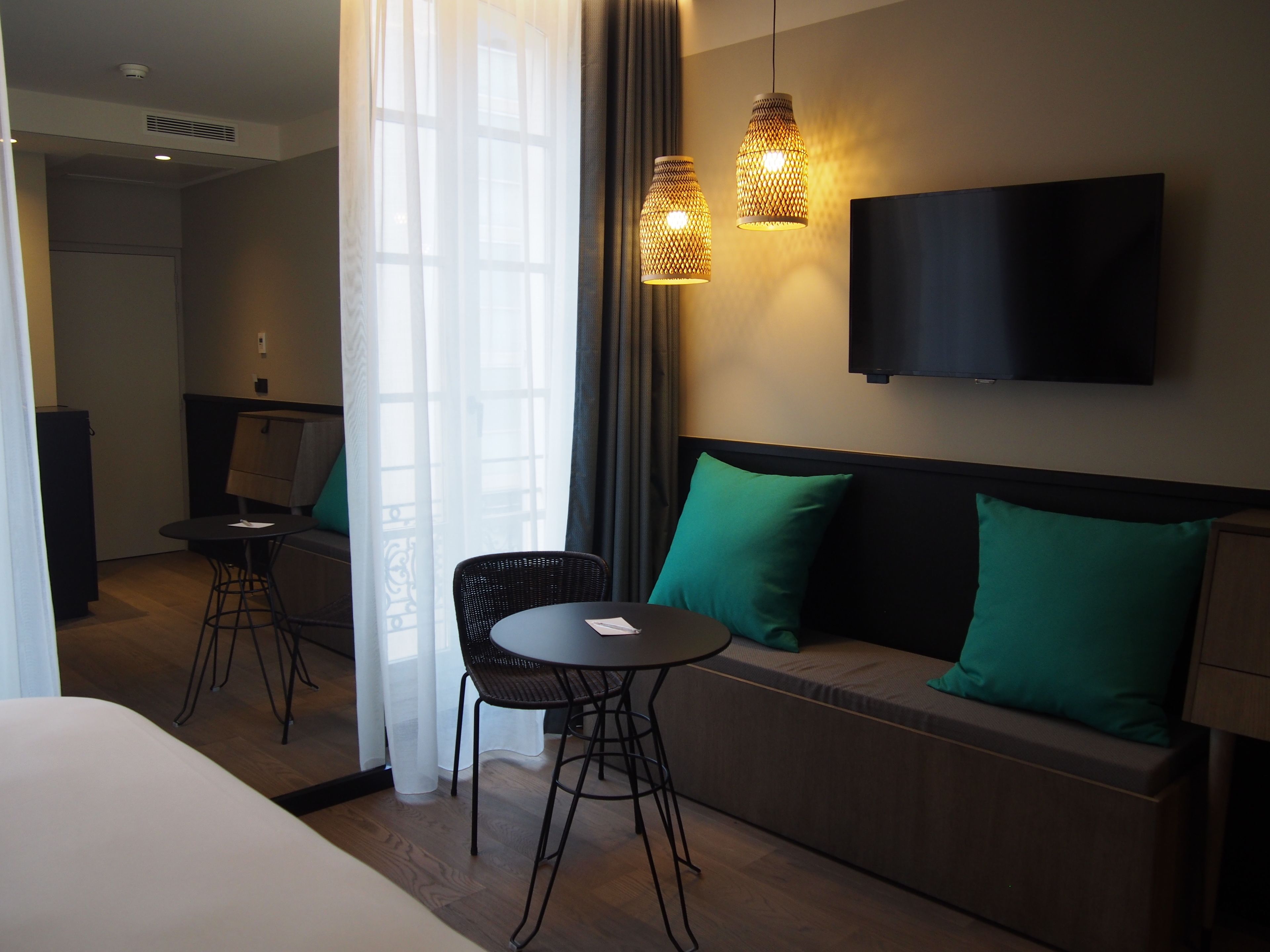 Habitación de hotel con cama, sofá con cojines verdes, una pequeña mesa con dos sillas, televisor montado en la pared y ventana con cortinas traslúcidas.
