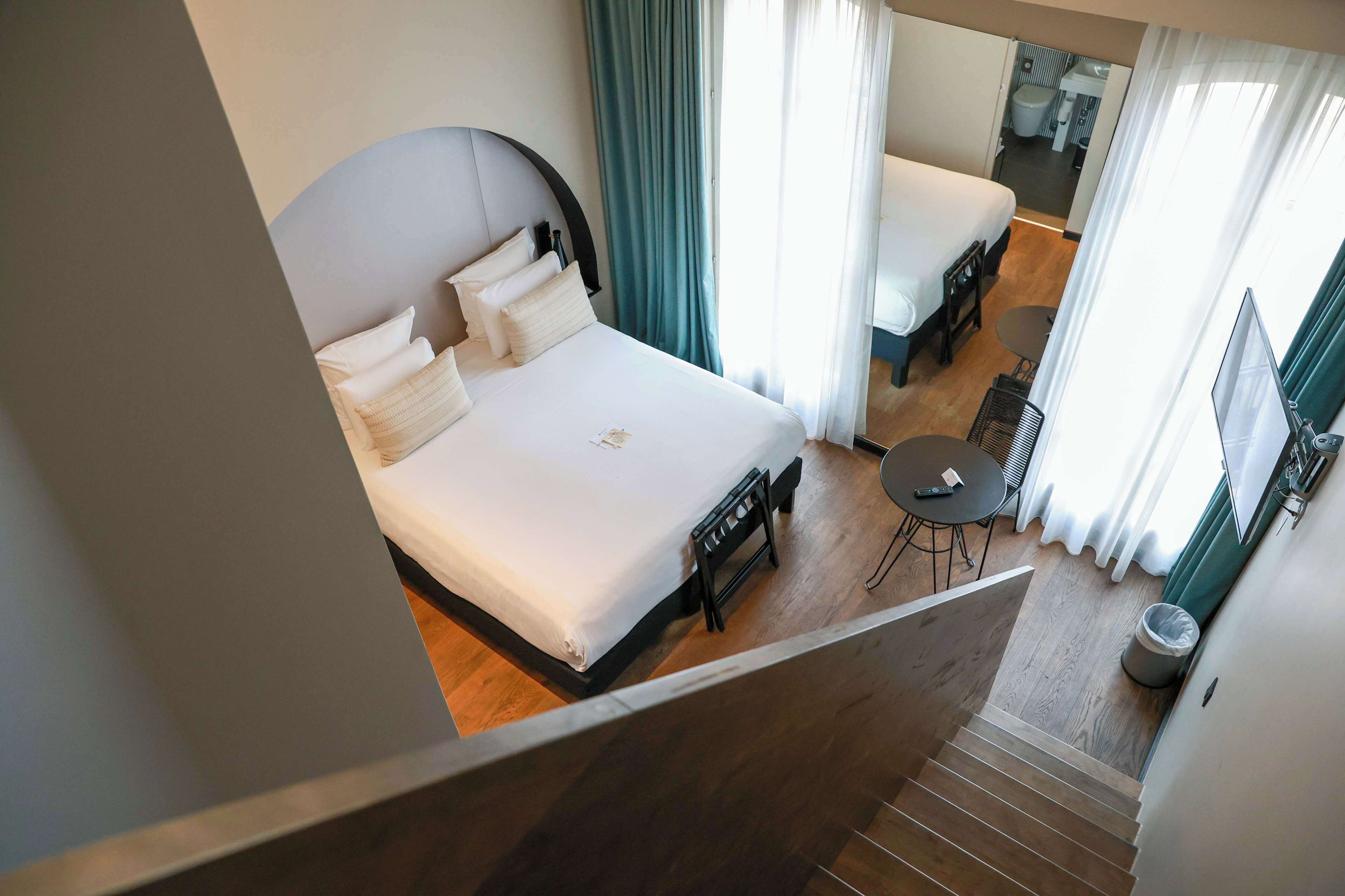 A hotel room with a large bed, a small desk, and a chair. There is a window with curtains, a trash can, and a bathroom visible in the background.