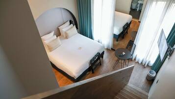 A hotel room with a large bed, a small desk, and a chair. There is a window with curtains, a trash can, and a bathroom visible in the background.
