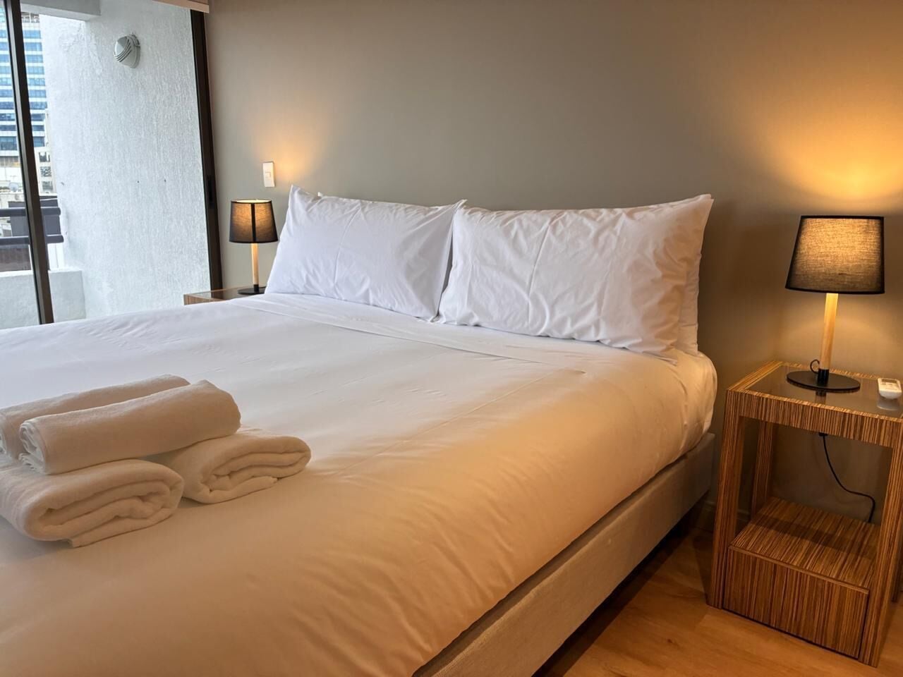 A hotel room with a large bed, two bedside lamps, a nightstand, and a window with a view of buildings.