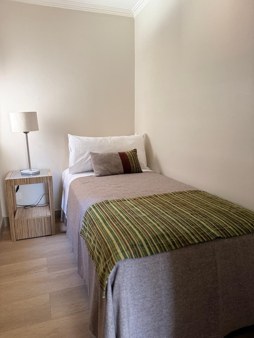 Una cama individual con una colcha a rayas, una lámpara de noche y una pequeña mesita de noche de madera.