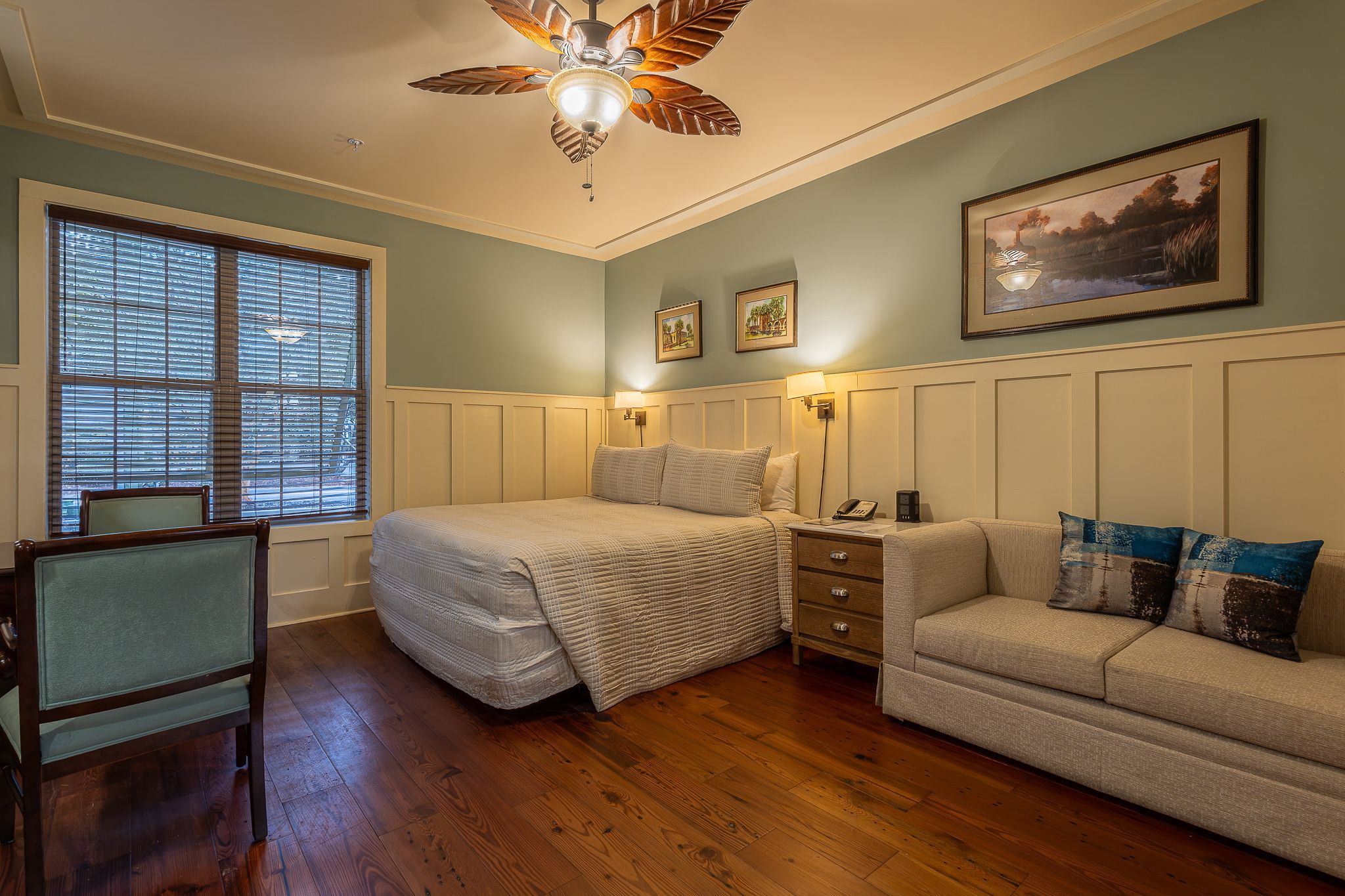 A bedroom with a bed, a nightstand, a dresser, a chair, a sofa, a ceiling fan, and a painting on the wall.
