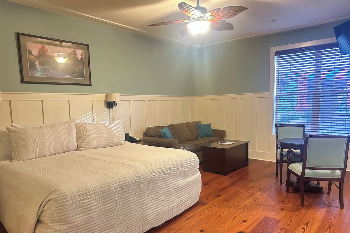 A bedroom with a large bed, a desk, a chair, a ceiling fan, a window with blinds, and a framed picture on the wall.