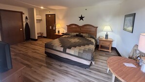 A hotel room with a wooden floor, a bed with a quilt, a desk with a lamp, a chair, a door, and a star decoration on the wall.