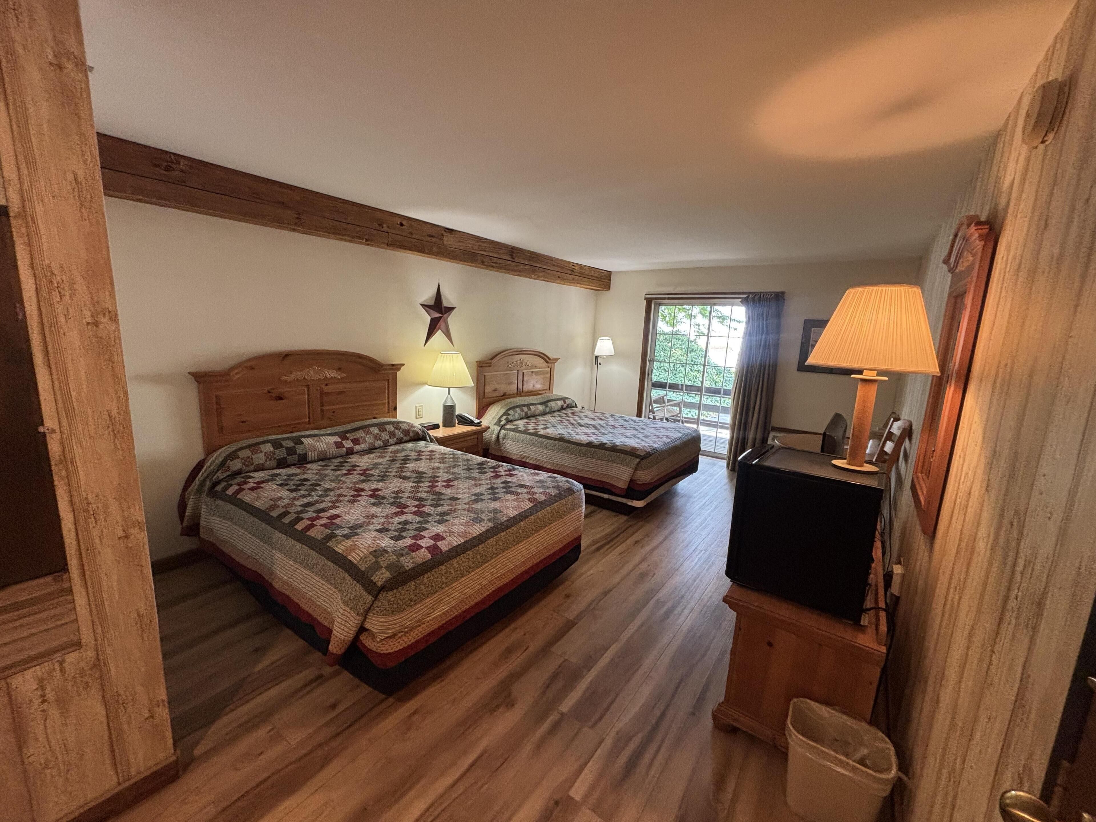 A room with two wooden beds, a star decoration on the wall, a desk with a lamp, and a view of the outdoors through a window.