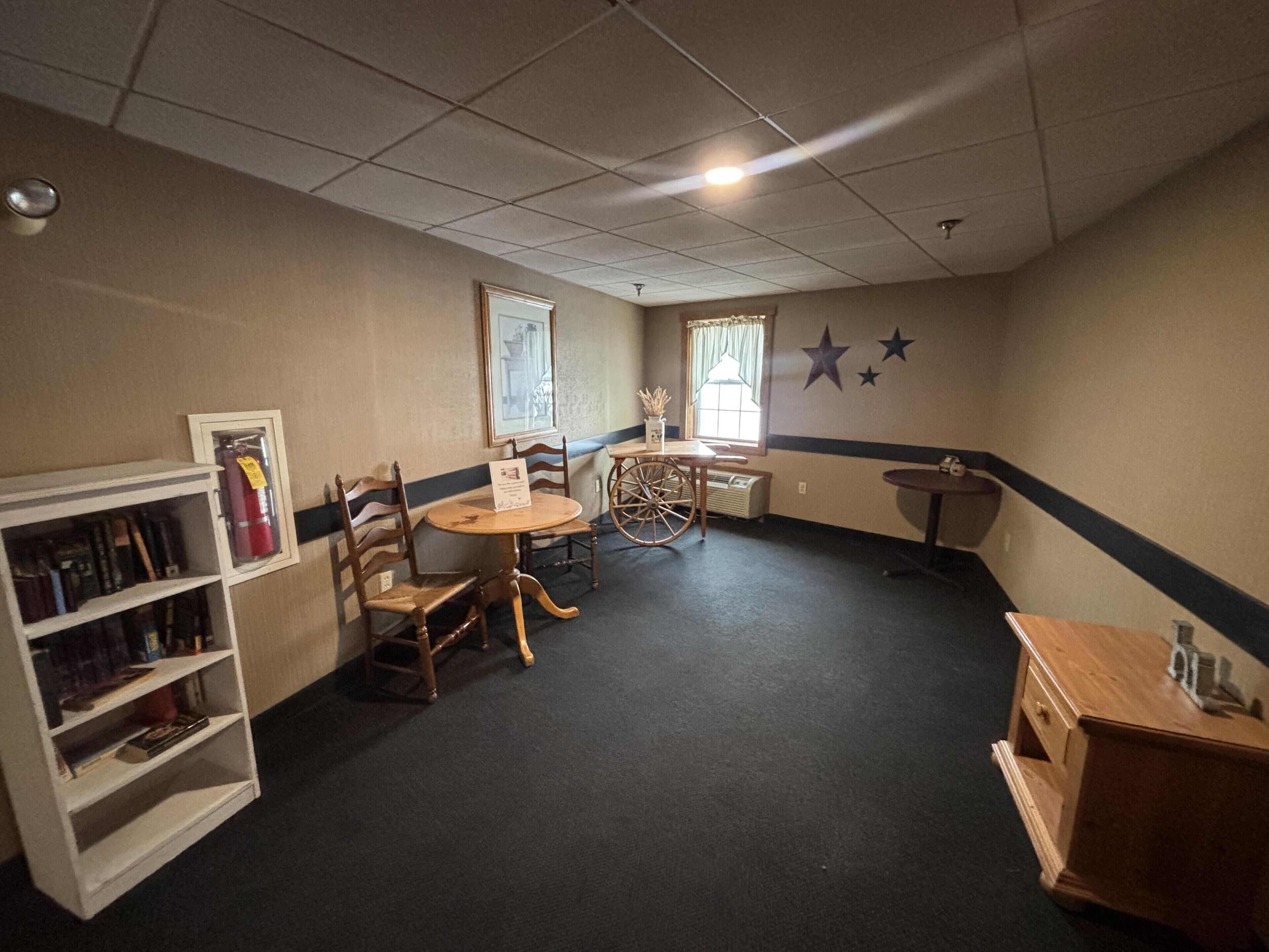 A room with a round table, two chairs, a bookshelf, a small wooden cabinet, and a window with curtains.