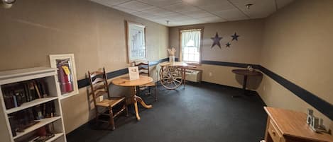 A room with a round table, two chairs, a bookshelf, a small wooden cabinet, and a window with curtains.