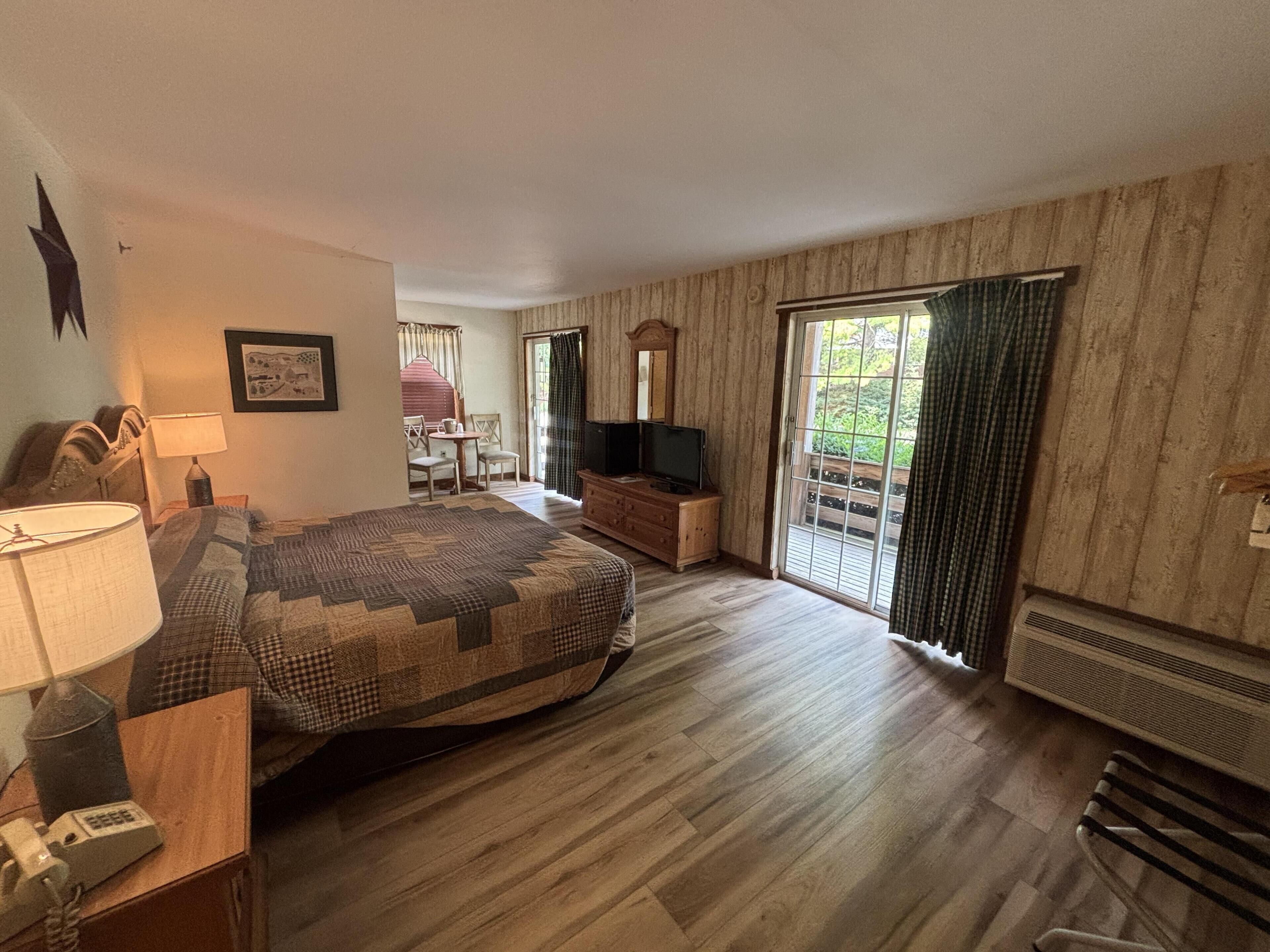 Quarto de hotel com cama, escrivaninha com luminária, cadeira, televisão e porta de vidro deslizante para uma varanda.