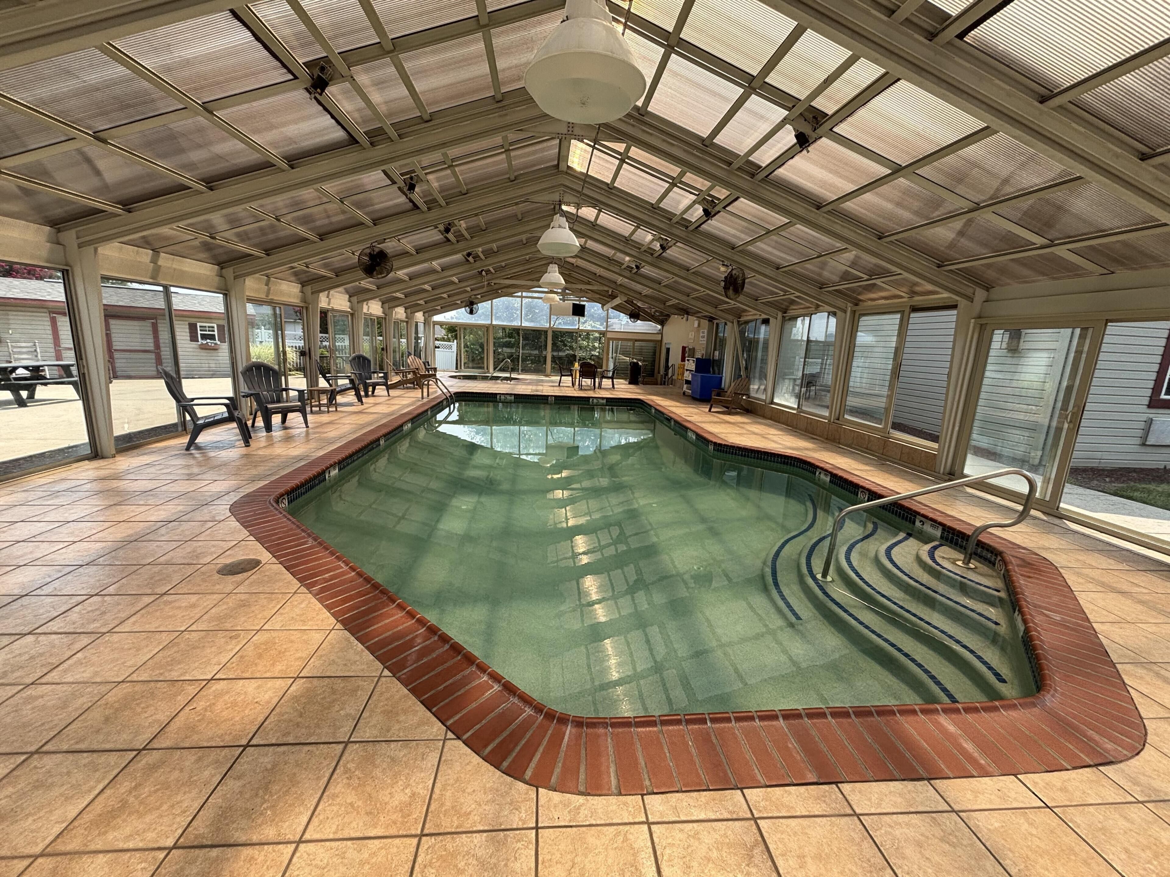 A covered swimming pool with a tiled floor, a poolside area with chairs, and a view through large windows.