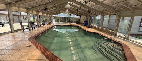 Una piscina techada con piso de baldosas, zona junto a la piscina con sillas y vistas a través de amplios ventanales.